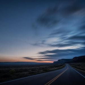 An empty two-lane highway curving into the distance at dusk, under a wide blue sky streaked with dark clouds and a band of orange light on the horizon.